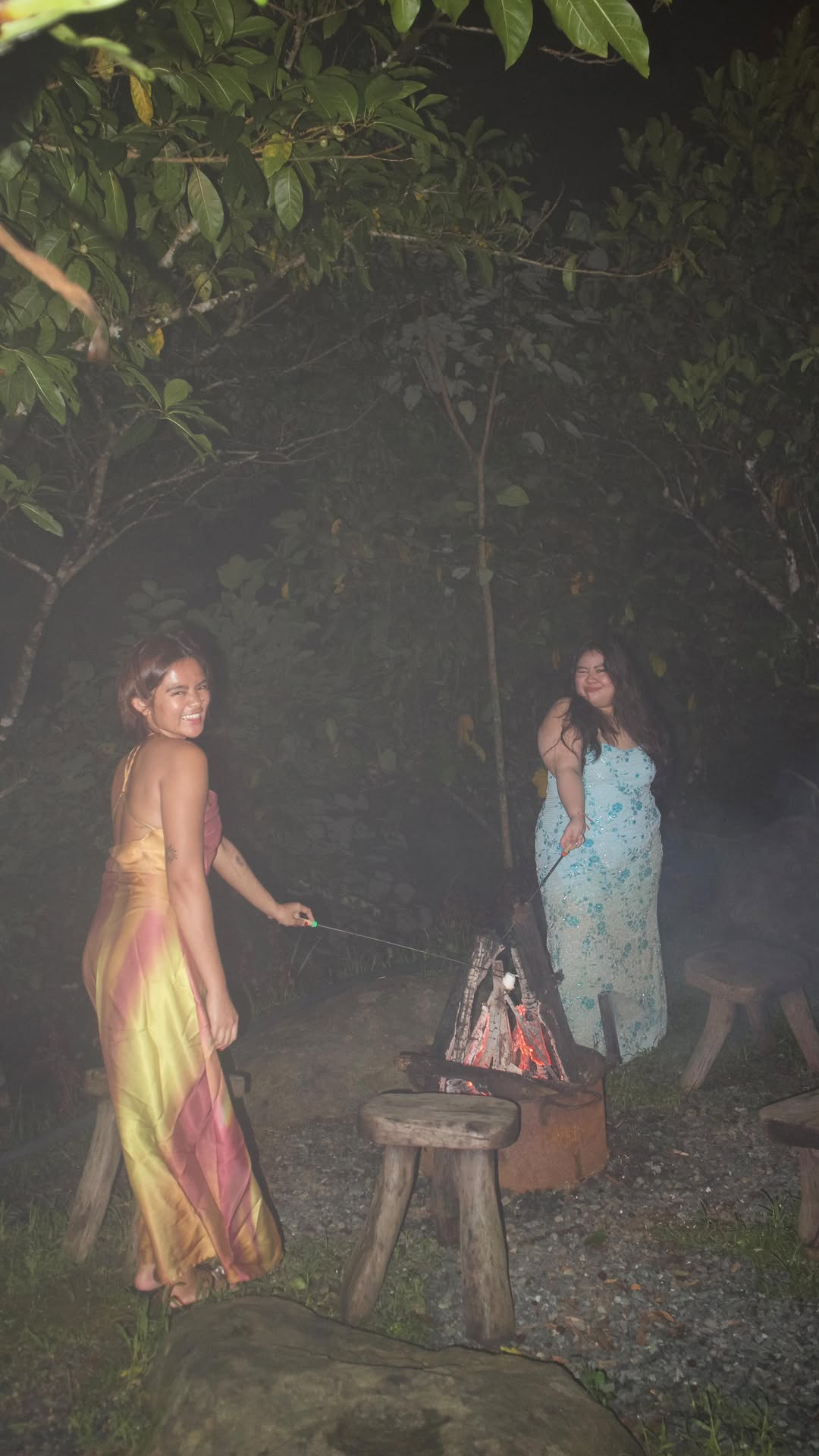 Two women in formal dresses laughing and roasting marshmallows around the La Cabaña bonfire at night